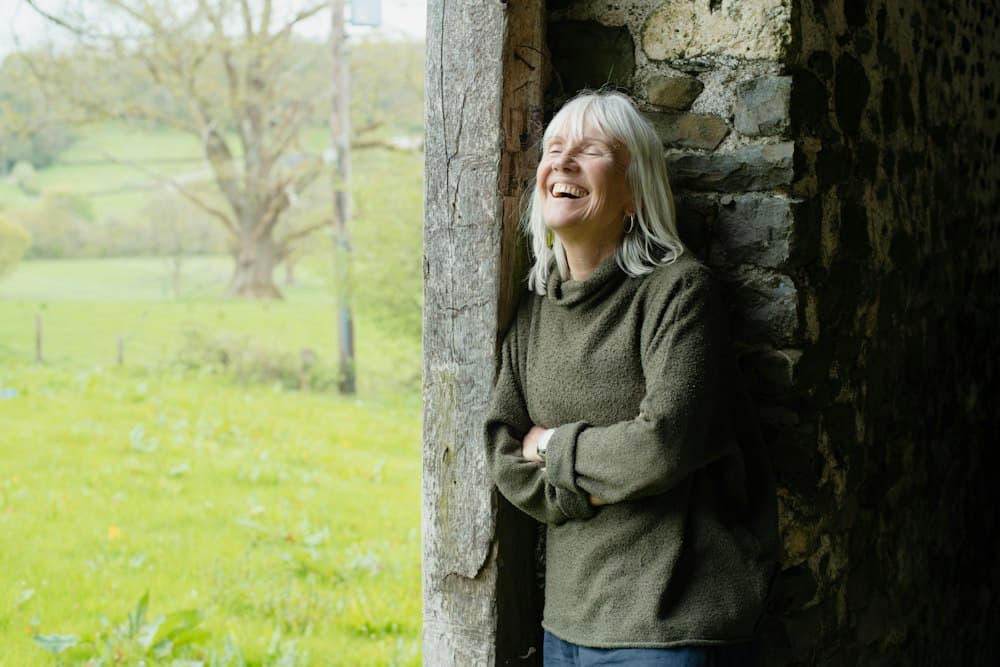 Femme aux cheveux blancs qui rit, appuyée à une porte ouverte sur la campagne