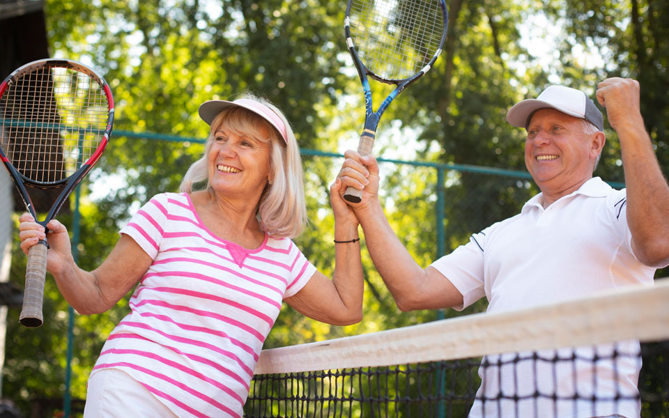 Deux seniors jouant au tennis sur un court extérieur, souriant et échangeant des balles.